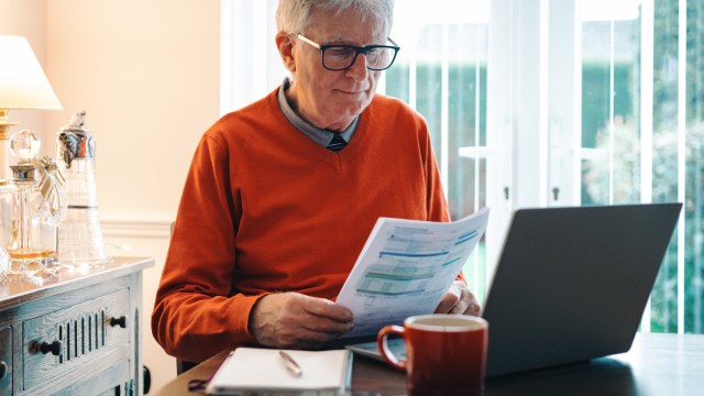 Article thumbnail: Portrait of a senior caucasian man in his 70s checking his energy bills at home. He has a worried expression while looking at the bills. Focus on the man while the interior architecture of the house is defocused.